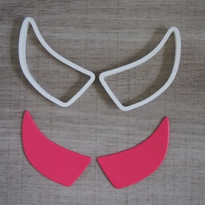 Op de afbeelding: Twee witte plastic koekjesvormen in de vorm van duivelshoorns en twee roze koekjesvormen in de vorm van duivelshoorns.