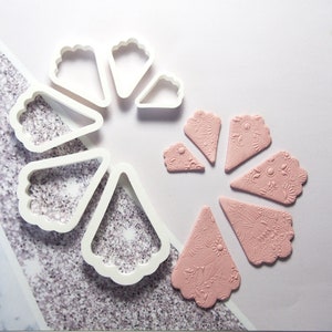 May include: A set of seven white plastic cookie cutters shaped like a flower with a scalloped edge. The cutters are arranged in a circle. Next to the cutters are seven pink fondant pieces that have been cut out using the cutters. The fondant pieces have a delicate floral design.
