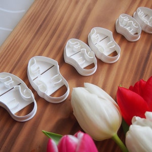 May include: Five white plastic cookie cutters shaped like baby shoes. The cutters are arranged in a row on a wooden surface. There are also some tulips in the background.