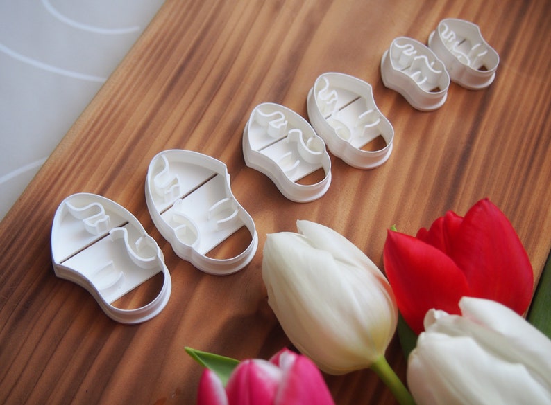 May include: Five white plastic cookie cutters shaped like baby shoes. The cutters are arranged in a row on a wooden surface. There are also some tulips in the background.