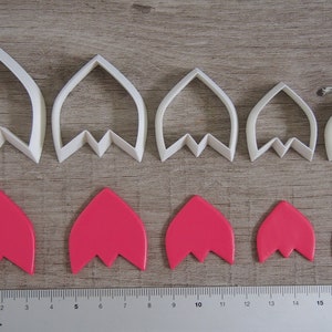 Peut inclure: Ensemble de six emporte-pi&egrave;ces &agrave; fleurs en plastique blanc de diff&eacute;rentes tailles, avec six formes de fleurs en p&acirc;te &agrave; sucre rose d&eacute;coup&eacute;es avec les emporte-pi&egrave;ces. Les emporte-pi&egrave;ces sont dispos&eacute;s en rang&eacute;e du plus grand au plus petit, et les formes en p&acirc;te &agrave; sucre sont dispos&eacute;es en rang&eacute;e sous les emporte-pi&egrave;ces. Une r&egrave;gle est visible en bas de l'image, avec le texte "Paper Poetry" imprim&eacute; dessus.