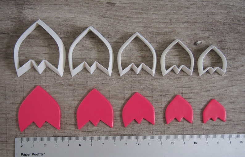 Peut inclure: Ensemble de six emporte-pi&egrave;ces &agrave; fleurs en plastique blanc de diff&eacute;rentes tailles, avec six formes de fleurs en p&acirc;te &agrave; sucre rose d&eacute;coup&eacute;es avec les emporte-pi&egrave;ces. Les emporte-pi&egrave;ces sont dispos&eacute;s en rang&eacute;e du plus grand au plus petit, et les formes en p&acirc;te &agrave; sucre sont dispos&eacute;es en rang&eacute;e sous les emporte-pi&egrave;ces. Une r&egrave;gle est visible en bas de l'image, avec le texte "Paper Poetry" imprim&eacute; dessus.