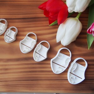 May include: A set of six white plastic cookie cutters shaped like faces with different features. The cutters are arranged in a row on a wooden surface.