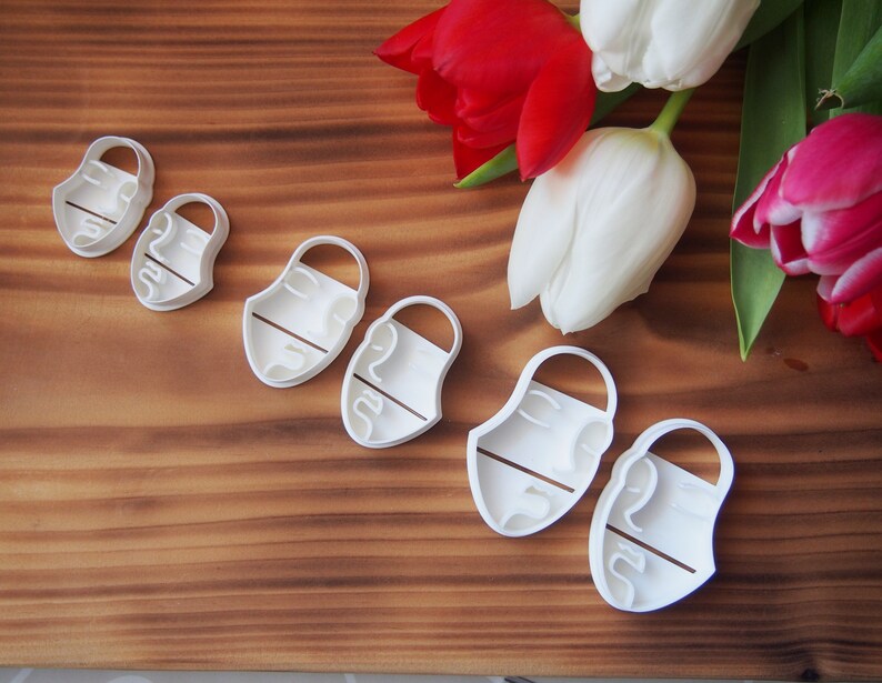 May include: A set of six white plastic cookie cutters shaped like faces with different features. The cutters are arranged in a row on a wooden surface.
