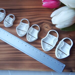 May include: A set of six white plastic cookie cutters shaped like abstract faces. The cutters are arranged in a row on a wooden surface. A ruler is visible in the background.