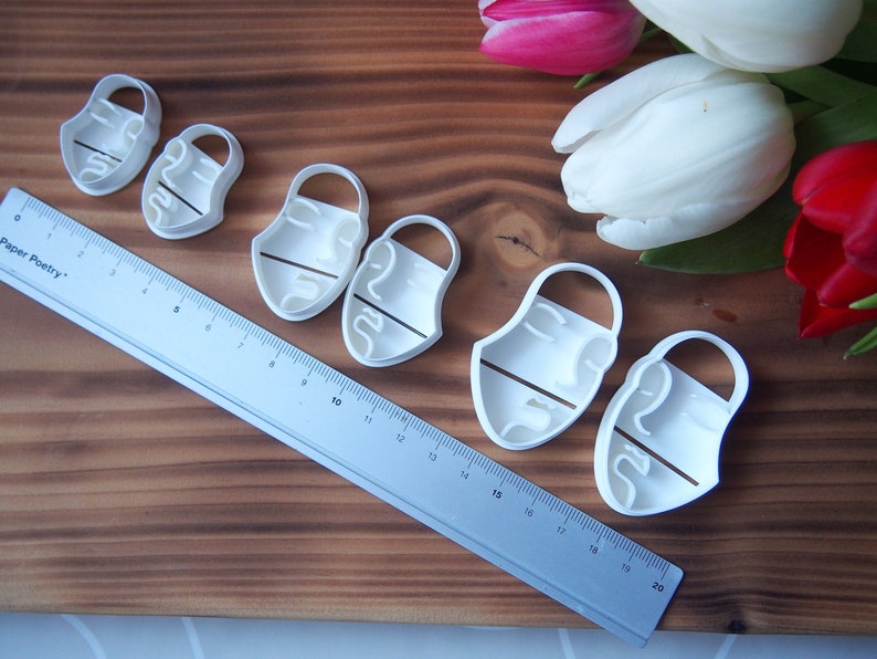 May include: A set of six white plastic cookie cutters shaped like abstract faces. The cutters are arranged in a row on a wooden surface. A ruler is visible in the background.