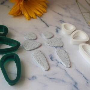 May include: A set of green and white polymer clay earrings. The green earrings are shaped like teardrops and the white earrings are shaped like irregular ovals with a speckled texture.