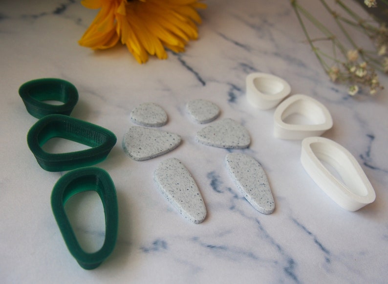 May include: A set of green and white polymer clay earrings. The green earrings are shaped like teardrops and the white earrings are shaped like irregular ovals with a speckled texture.