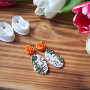 May include: A pair of polymer clay earrings with orange flower accents and a leaf design. The earrings are on a wooden surface with white and pink tulips.