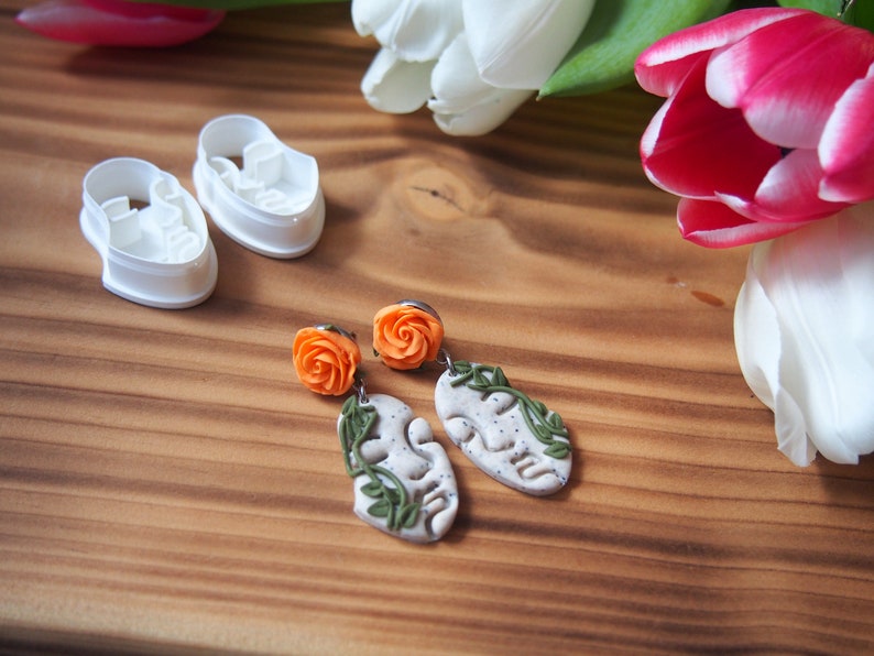 May include: A pair of polymer clay earrings with orange flower accents and a leaf design. The earrings are on a wooden surface with white and pink tulips.