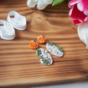 May include: A pair of handmade earrings featuring a white ceramic leaf design with a green vine and an orange flower charm.