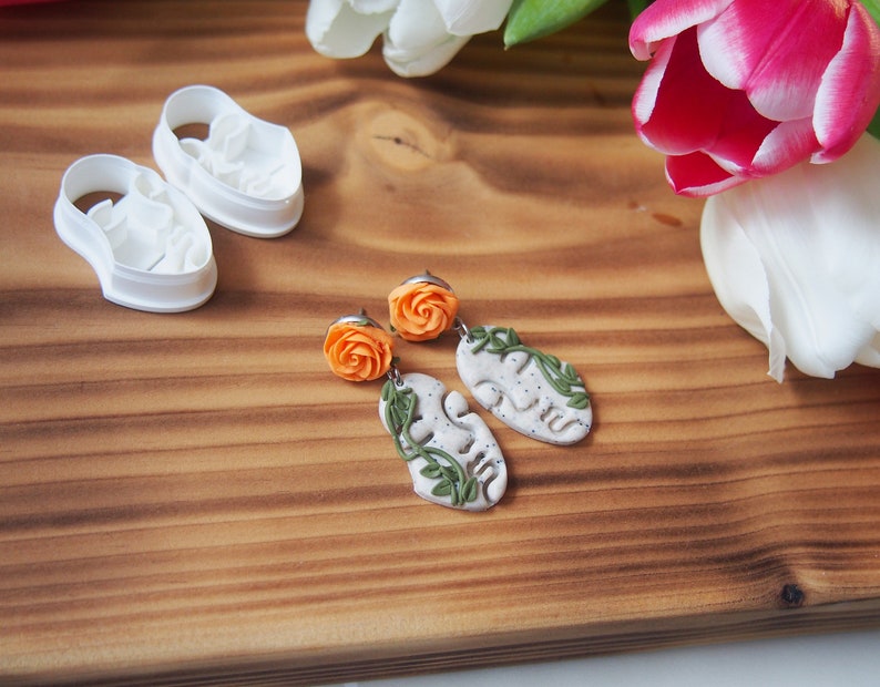 May include: A pair of handmade earrings featuring a white ceramic leaf design with a green vine and an orange flower charm.