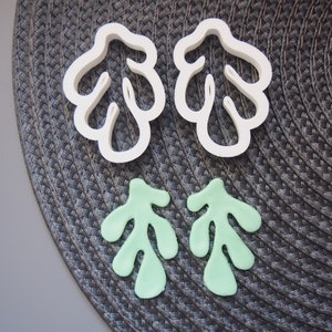 May include: Two white plastic cookie cutters shaped like a leaf with a jagged edge. Two green leaf-shaped cookies are also shown.