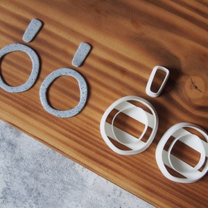 May include: A set of five cookie cutters in various shapes and sizes. The cutters are made of white plastic and feature a speckled grey finish. The set includes two large oval-shaped cutters, one small oval-shaped cutter, and two rectangular cutters.