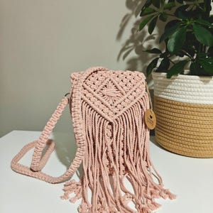 May include: A light pink macrame crossbody bag with a geometric design on the flap and long fringe. The bag has a long, adjustable strap and a small wooden tag that says "Love". The bag is sitting on a white surface.
