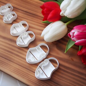 May include: Set of six white plastic cookie cutters shaped like tulips. The cutters are arranged in a row on a wooden surface. There are also several tulips in the background, some white and some red.