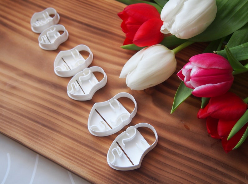 May include: Set of six white plastic cookie cutters shaped like tulips. The cutters are arranged in a row on a wooden surface. There are also several tulips in the background, some white and some red.