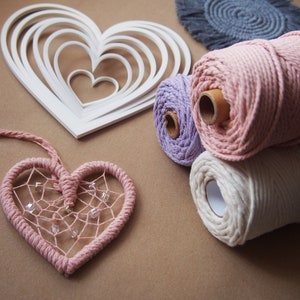 Peut inclure: Trois bobines de cordon en coton rose, violet et blanc. Le cordon est utilisé pour le macramé et d'autres travaux manuels. Un gabarit en forme de cœur blanc et un attrape-rêves en forme de cœur rose sont également présents sur l'image.