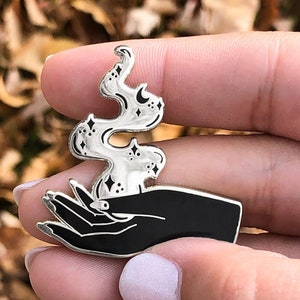 May include: A silver enamel pin featuring a black hand holding a silver flame with stars and crescent moons.