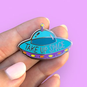May include: A silver and blue enamel pin shaped like a UFO with the text "Take Up Space" in purple and yellow lettering.