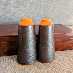 May include: A pair of ceramic salt and pepper shakers. The shakers are dark brown with a textured finish and orange tops. The bases are also orange. The shakers are displayed on a wooden surface.