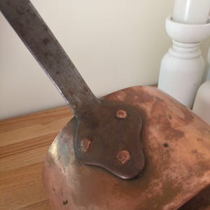 May include: Close-up of a copper saucepan with a long, riveted metal handle. The pan has a rounded shape and a dark, aged patina. The handle is attached with three rivets. The background is a light wood surface.