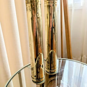 Op de afbeelding: Twee hoge, cilindrische messing vazen met een gepolijste afwerking staan op een spiegeltafel. De vazen hebben een licht verouderd uiterlijk, met wat zichtbare slijtage. De achtergrond omvat een transparant gordijn en een houten driepootlamp.
