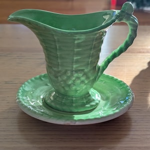 May include: A green ceramic creamer and saucer set. The creamer has a basketweave texture and a curved handle. The saucer is round with a matching texture. The set is a light green color and sits on a wooden surface.