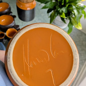Puede incluir: Primer plano de la base de una olla de cerámica con un esmalte naranja óxido. La olla tiene un borde blanco y una firma manuscrita y el número 7. Otras piezas de cerámica y una planta están en el fondo.
