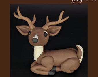 May include: A handcrafted ceramic deer figurine in shades of brown, with a white chest and a white patch on its rear. The deer has large, dark eyes, a black nose, and detailed antlers. The figurine is in a resting position.