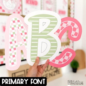 May include: Large, decorative ABC letters in pink, green, and white. The letter A has a pink and white checkered pattern with small hearts. The letter B is green with white stripes. The letter C has a pink and white checkered pattern. The text "PRIMARY FONT" is at the bottom.