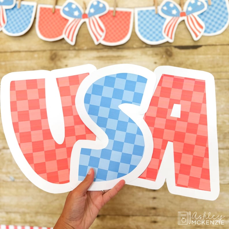 Op de afbeelding: Het woord "USA" in grote, uitgesneden letters, met een rood-blauw geblokt patroon. De letters worden door een hand vastgehouden, met een houten achtergrond. Een banner met patriottische strikken en vormen hangt erboven.