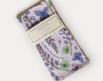 May include: A light purple cotton tea towel featuring a botanical print of blue butterflies and purple lupine flowers. A paper band across the center reads Cotton Tea Towel, 19 by 28 inches, hang loop, Made in the USA, CLAIRE LOON BALDWIN.