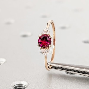 May include: A rose gold ring featuring a round, deep red ruby gemstone surrounded by smaller, white diamonds.