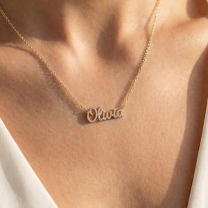 Könnte beinhalten: Eine goldfarbene Halskette mit dem Namen "Olivia" in Schreibschrift. Die Halskette wird um den Hals getragen und liegt auf einem weißen Oberteil. Der Name ist in einer zarten Schriftart gehalten.