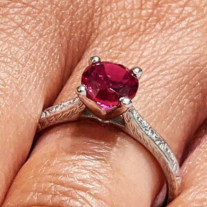 May include: A silver ring with a red gemstone set in the centre. The ring has a delicate, textured band.