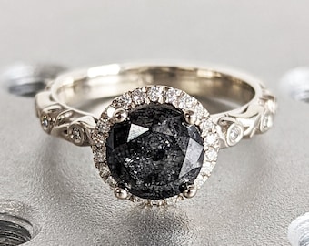 May include: A silver engagement ring with a black diamond set in a halo of smaller diamonds. The ring is on a gray textured surface.