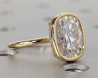 May include: A gold ring with a large, clear, cushion-cut gemstone set in a bezel setting. The ring is on a gray, textured surface.