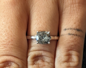 May include: A silver ring with a large, square-shaped, grey gemstone set in a prong setting. The ring is on a person's finger.
