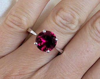 May include: A silver ring with a large, round, deep red gemstone. The gemstone is the focal point, set in a simple, elegant band. The ring is shown on a hand with light pink nail polish.