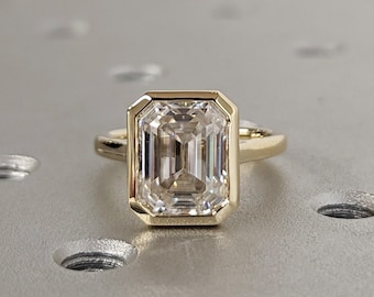 Könnte beinhalten: Ein goldener Ring mit einem Smaragdschliff-Diamanten. Der Ring liegt auf einer grauen Oberfläche mit kleinen Löchern.