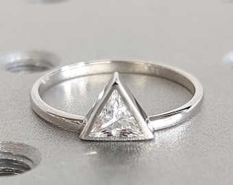 May include: A silver ring with a triangular, clear gemstone. The ring's band is thin and polished, and the gemstone is set in a bezel setting. The ring is displayed on a textured, light gray surface.
