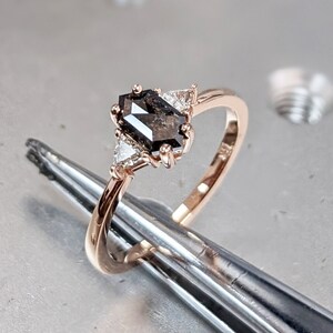 May include: A rose gold engagement ring with a black diamond center stone and two smaller diamonds on the side. The ring is held by tweezers on a gray surface.
