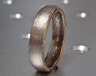 May include: A silver and gold ring with a textured pattern. The ring is on a gray surface with small holes.