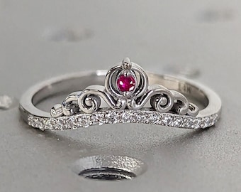 May include: A silver ring with a crown design and a small red gemstone in the center. The band is decorated with small clear gemstones.
