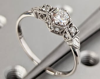 May include: A silver ring with a round, clear diamond set in a delicate, ornate design. The ring is on a gray surface with small holes.