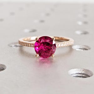 May include: A rose gold ring featuring a large, round, bright pink gemstone. The band is embellished with small, sparkling accent stones. The ring is set on a light, textured surface, showcasing its elegant design.