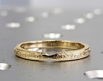 Könnte beinhalten: Ein goldener Ehering mit einem komplizierten gravierten Design. Der Ring ist dünn und hat eine leicht abgerundete Form.