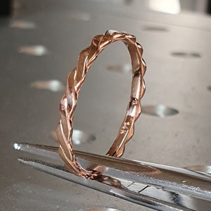 May include: A rose gold band ring with a twisted design. The ring is held by tweezers on a metal surface.