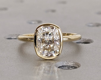 Könnte beinhalten: Ein goldener Ring mit einem großen, klaren Diamanten im Kissen-Schliff, der in einer Zargenfassung sitzt.
